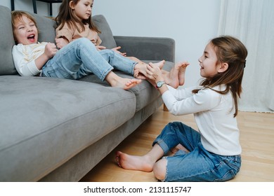 Side View Of A Girl Conducting Tickling Competition Between Siblings Sitting On A Couch Barefoot.