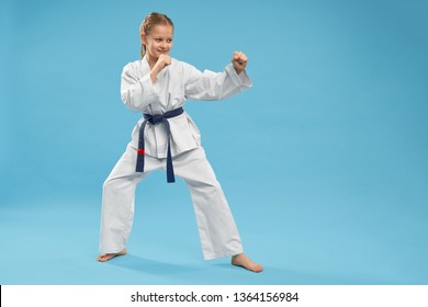 Side View Of Girl Doing Martial Arts On Blue Isolated Background. Active Teenager In White Kimono Looking At Opponent And Fighting In Studio. Child Doing Sport And Exercising. Concept Of Combat. 