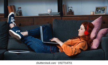 Side view of girl in headphones using laptop near digital tablet on couch  - Powered by Shutterstock