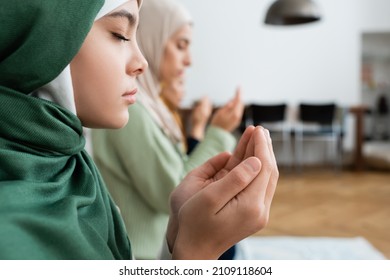 Side view of girl in hijab praying near blurred family at home