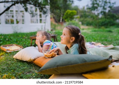 Side view of girls lying on blanket, eating homemade popcorn and watching film on DIY screen from projector. Summer outdoor weekend activities with kids. Open air cinema. Outside movie night