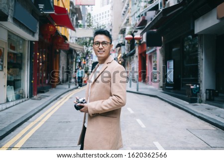 Side view of glad Asian male tourist in trendy beige greatcoat and stylish eyeglasses photographing with camera on street road and looking away  