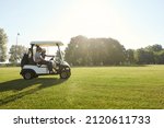 Side view of golfers riding golf cart on green lawn of golf field at warm sunny day. Concept of entertainment, recreation, leisure and hobby outdoors. Idea of frienship. Young caucasian male friends