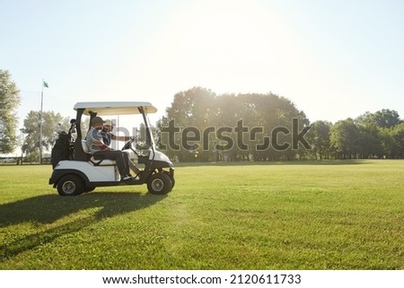 Side view of golfers riding golf cart on green lawn of golf field at warm sunny day. Concept of entertainment, recreation, leisure and hobby outdoors. Idea of frienship. Young caucasian male friends