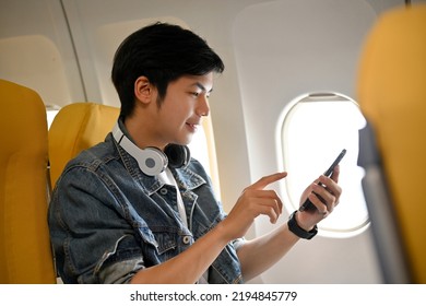 Side View, A Good Looking Asian Male Traveler In Jean Jacket And Headphones Sits At The Window Seat In Economy Class, Using His Mobile Phone To Check His Email Before Takeoff. Airplane Journey Concept
