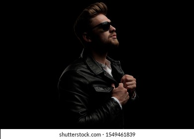 side view of a gorgeous casual man wearing leather jacket standing and fixing his jacket with confidence on black studio background