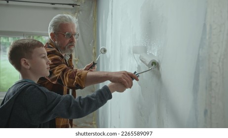 Side view of grandfather and grandson painting wall with white paint using paint rollers. An elderly man teaching a young guy and giving advice. The concept of family, teamwork, apartment renovation. - Powered by Shutterstock