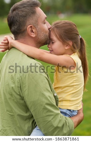 Side view of grateful dad holding and kissing his little girl while spending time together outdoors on a warm day