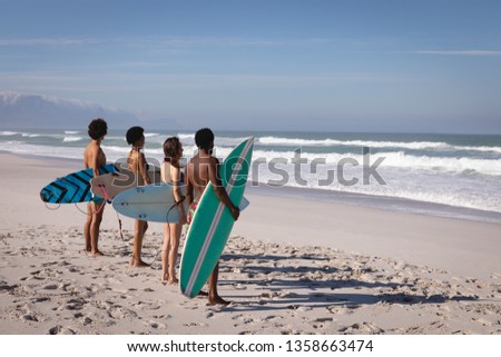 Side view of group of multi-ethnic friends standing in front of the sea with their surfboards and looking at the waves at beach in the sunshine