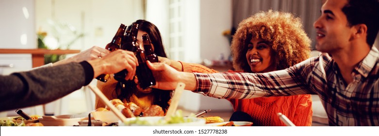 Side View Of A Group Of Young Adult Multi-ethnic Male And Female Friends Sitting At A Table At Home Set For Thanksgiving Dinner Making A Toast With Bottles Of Beer