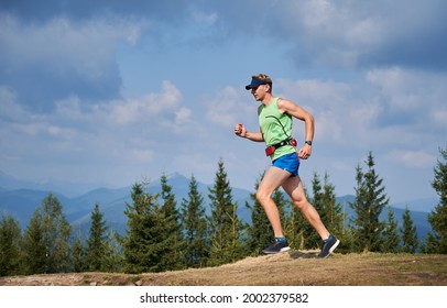 Side View Of Guy Running In Fresh Air By Mountain Trails. Summer Trail Running Marathon In Mountains On Background Of Green Trees, Cloudy Sky And Silhouettes Of Mountain Beskids.