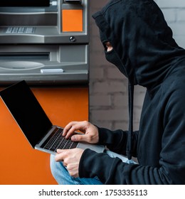 Side View Of Hacker In Balaclava Using Laptop Near Automated Teller Machine