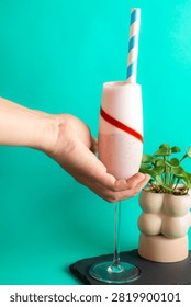 Side view of a hand holding and presenting an elegant glass of fruity smoothie with a straw over a bright colorful background.