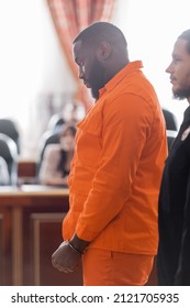 Side View Of Handcuffed African American Man In Jail Uniform Near Bailiff And Blurred Jurors In Court