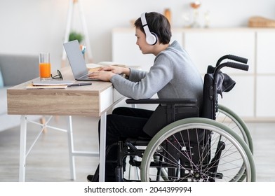 Side View Of Handicapped Teen Boy In Wheelchair Wearing Headphones, Having Video Chat With Teacher Or Classmate At Home. Disabled Teenager Studying Online, Having Remote Lesson