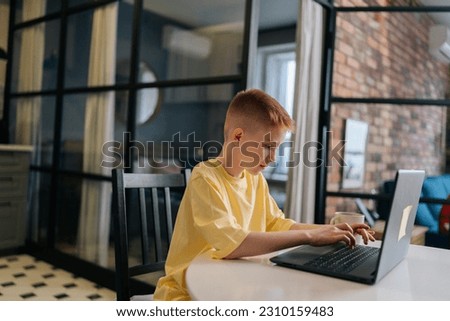 Side view of handsome 10 year old boy in yellow t-shirt typing on laptop keyboard during online lesson, distance studying. Focused child schoolboy using computer sitting at table in living room.