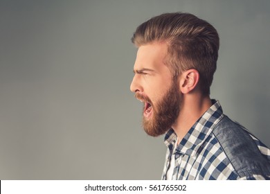 Side view of handsome bearded man in casual clothes screaming, on gray background