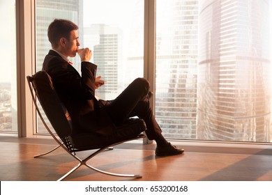 Side View Of Handsome Businessman Sitting In Comfortable Office Chair Enjoying Cup Of Coffee In The Morning, Looking Through Big Window At Dawn Sunrise City, Getting Motivation For New Working Day