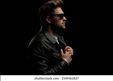 side view of a handsome casual man standing and fixing his jacket while looking ahead confident on black studio background