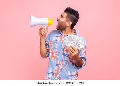 Side view of handsome crazy screaming in megaphone man with beard holding fan of dollars, wearing blue casual style shirt, announcing bonus. Indoor studio shot isolated on pink background.