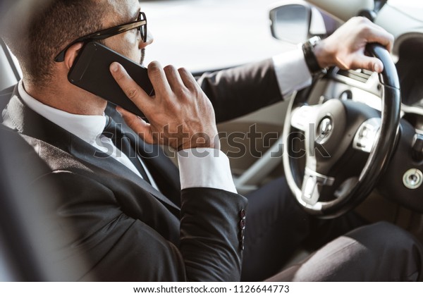 side view of handsome driver in suit driving\
car and talking by\
smartphone