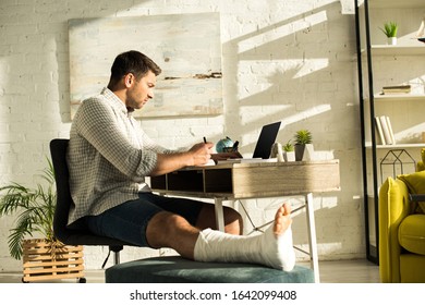 Side View Of Handsome Freelancer With Broken Leg Writing On Notebook And Using Laptop At Table