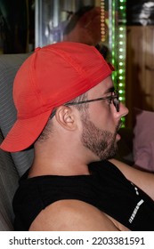 Side View Of A Handsome Guy With Red Hat, Glasses And Beard Looking At Something He Has In Front Of Him Inside The House