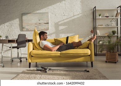 Side View Of Handsome Man With Broken Leg Using Laptop On Couch