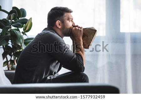 side view of handsome man with panic attack breathing in paper bag in apartment 