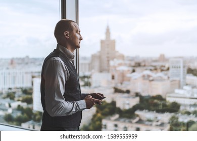 Side view of a handsome mature businessman pensively looking outside of a window on a top floor of his business office high-rise, a smartphone in his hands, with a copy space zone on the right