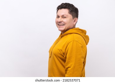 Side View Of Handsome Middle Aged Man With Dark Hair Standing Looking At Camera With Satisfied Face And Smiling, Wearing Urban Style Hoodie. Indoor Studio Shot Isolated On White Background.