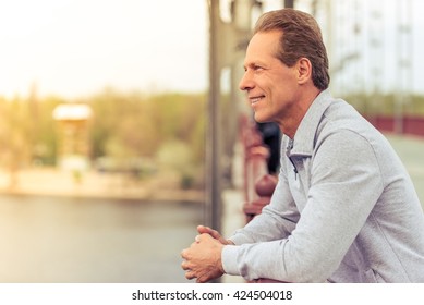 Side View Of Handsome Middle Aged Man In Sports Uniform Leaning On Bridge And Smiling While Having Break During Morning Run