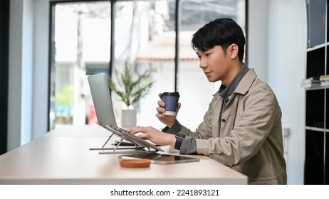 side view, Handsome and smart young Asian male programmer or freelance graphic designer remote working at the coffee shop, sipping coffee while using laptop computer. - Powered by Shutterstock