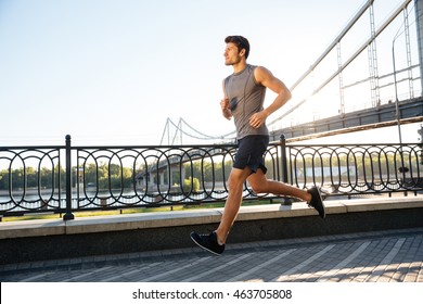 Side View Of A Handsome Sports Man Running Fast Along Big Modern Bridge At Sunset Light