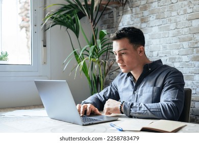 Side view handsome young businessman in eyewear working with computer remotely, sitting at wooden table in office. Pleasant happy man communicating in social network, searching information online. - Shutterstock ID 2258783479