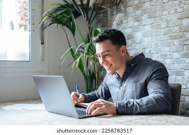 Side view handsome young businessman in eyewear working with computer remotely, sitting at wooden table in office. Pleasant happy man communicating in social network, searching information online. - Shutterstock ID 2258783517