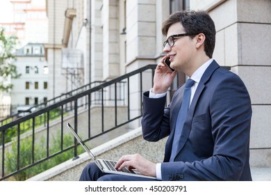 Side View Of Handsome Young Businessman Sitting Outside On Building Stairs, Using Laptop And Having Mobile Phone Conversation