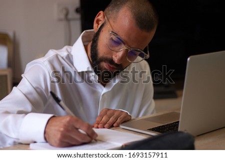 Side view handsome young businessman in eyewear working with computer remotely, sitting at wooden table. Pleasant happy man communicating in social network, searching information online.