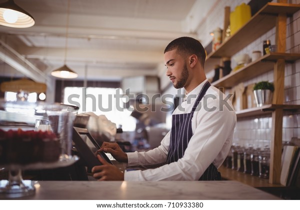 Side view of handsome young waiter using computer at\
counter in coffee shop