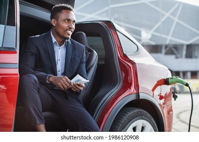 Side View Of Happy African America Man In Business Suit Counting Money Cash While Charging Electric Car. Young Man Sitting Inside Auto And Feeling Satisfaction About Economical Vehicle.