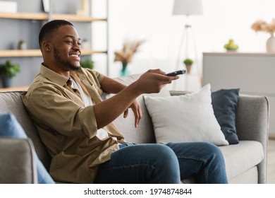 Side View Of Happy African American Man Watching TV Switching Television Channels With Remote Control Sitting On Sofa At Home. Weekend Leisure And Entertainment, TV Programming Concept