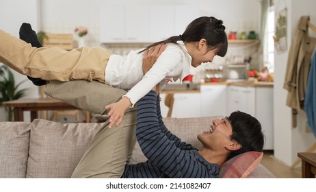 Side View Happy Asian Father Lying On Sofa And Holding Up Daughter On His Legs At Home. The Girl Open Arms And Flying In Air Like Plane