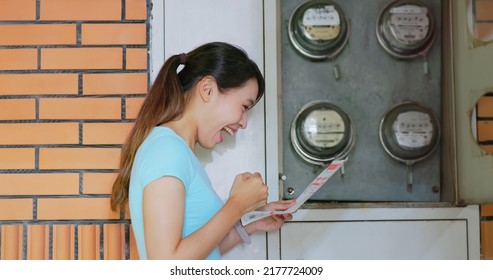 side view of happy asian woman stand by round electric meter on wall  smile happily posing fist gesture look at electricity bill saving in her hand