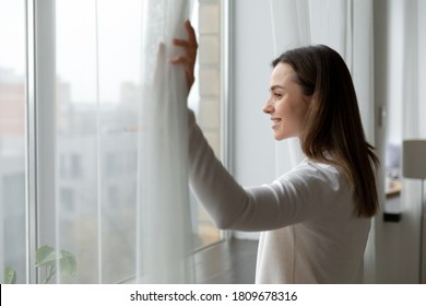 Side view happy attractive woman opening curtains, admiring cityscape at home or hotel room. Smiling millennial girl enjoying morning leisure weekend time or new day start, planning future indoors.