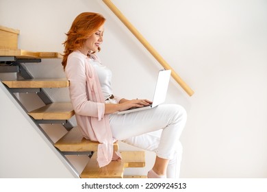 Side View Of Happy Beautiful Young Woman Working On A Laptop On Her Lap, Wearing Casual Clothes, While Sitting On The Stairs At Home