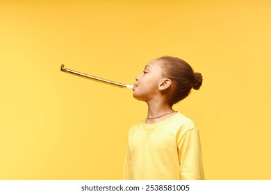 Side view of happy Black girl with cute hair bun blowing in party horn wearing yellow t shirt while standing against background of matching color in studio, copy space