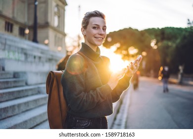 Side View Of Happy Blonde Female Student Using Mobile Phone While Looking At Camera Near Stairs Of Old Building In Sun Back Lit 