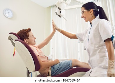 Side View Of Happy, Brave Kid Sitting In Chair In Dental Clinic Office And Giving High Five To Professional Female Therapist. Smiling Dentist Building Trust With Little Patient During Regular Check Up