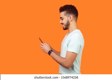 Side view of happy brunette man with beard in white t-shirt smiling while using cellphone, chatting in social media, enjoying mobile service. empty copy space for advertisement, indoor studio shot