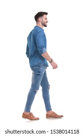 Side View Of A Happy Casual Man Walking Forward While Wearing Blue Shirt And Jeans On White Studio Background